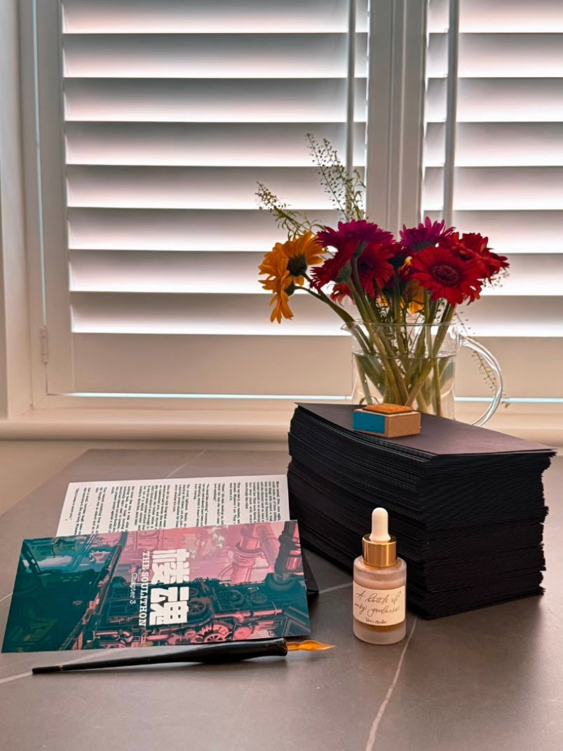 A styled desk scene with a stack of black notebooks, a small glass dropper bottle, an open book, a pen, and a vase of colorful flowers in front of white window blinds.